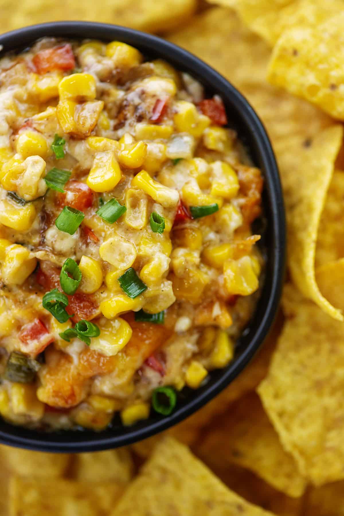 Overhead view of hot corn dip in black bowl surrounded by chips.