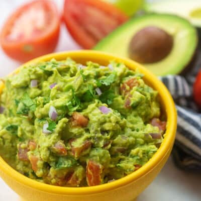 A close up of a bowl of Guacamole with sliced avocado in the background.