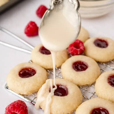 Spoonful of glaze being drizzled over thumbprint cookies on wire rack.