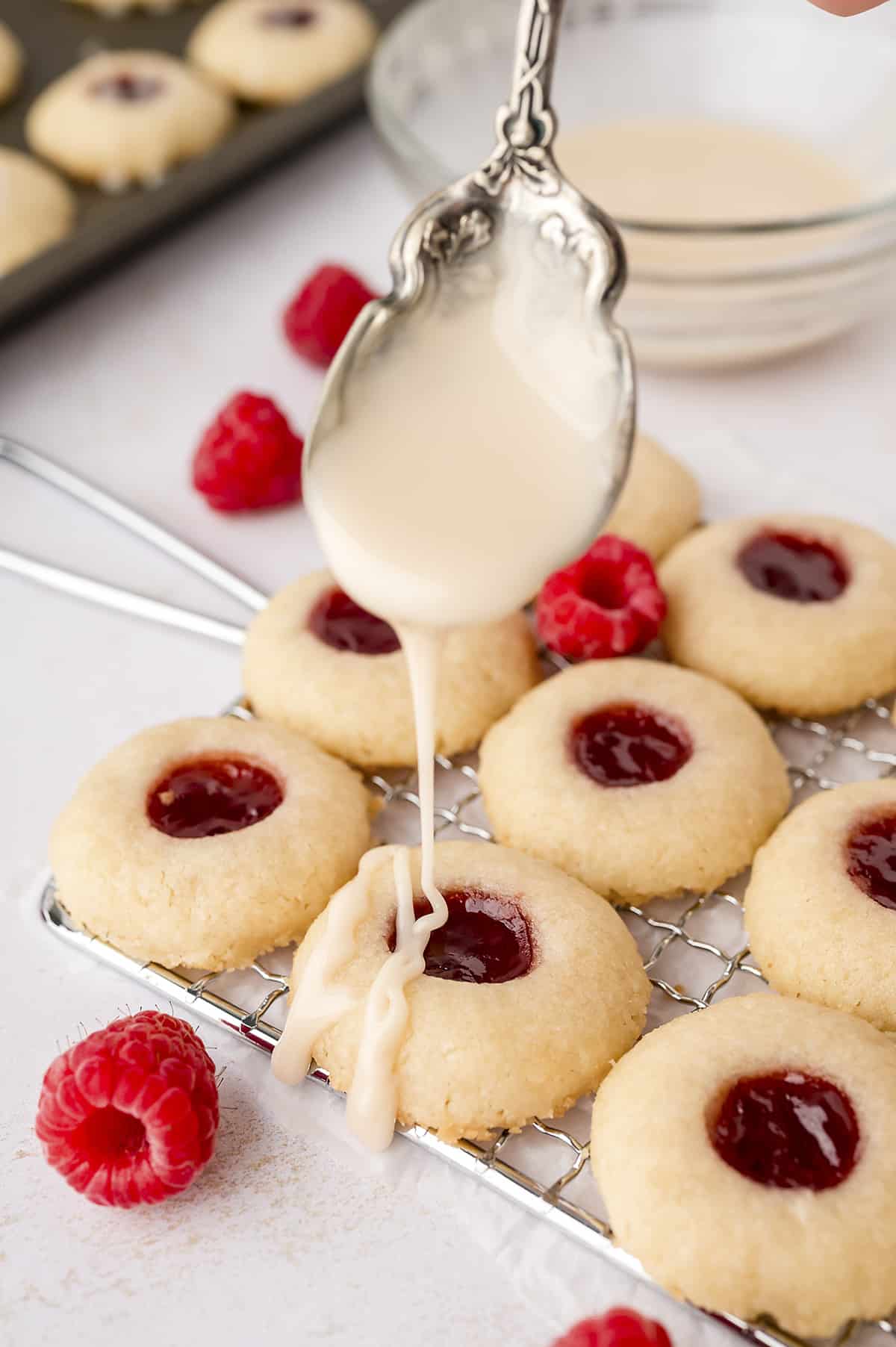Spoonful of glaze being drizzled over thumbprint cookies on wire rack.