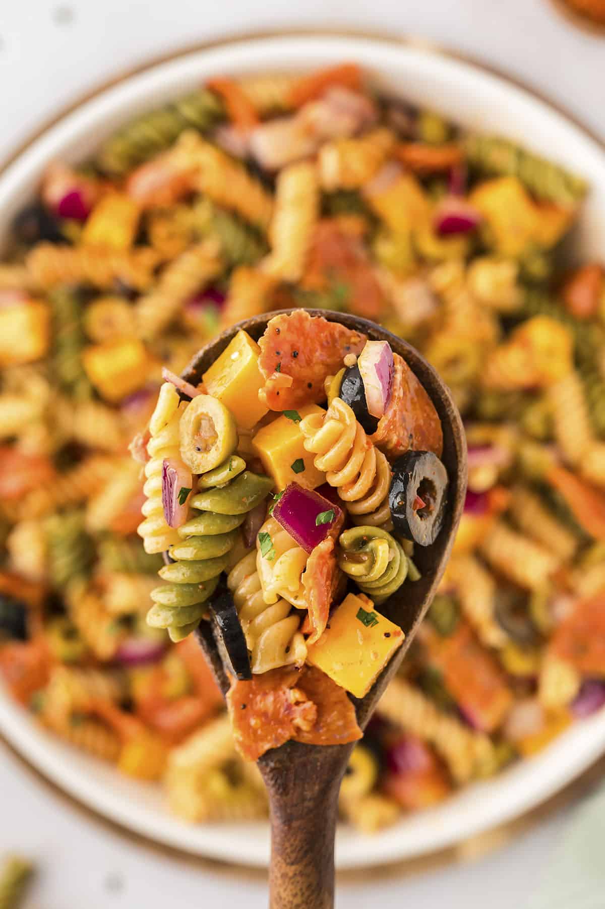 Cold pasta salad on wooden spoon over mixing bowl.