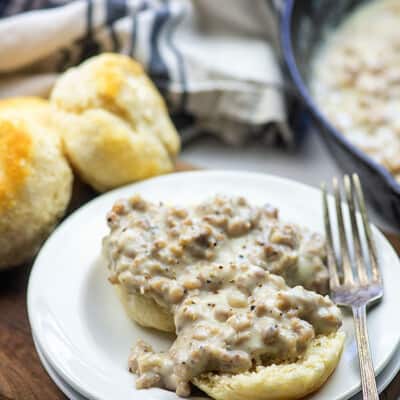 A single biscuit split in half topped with gravy in front of a cast-iron skillet.