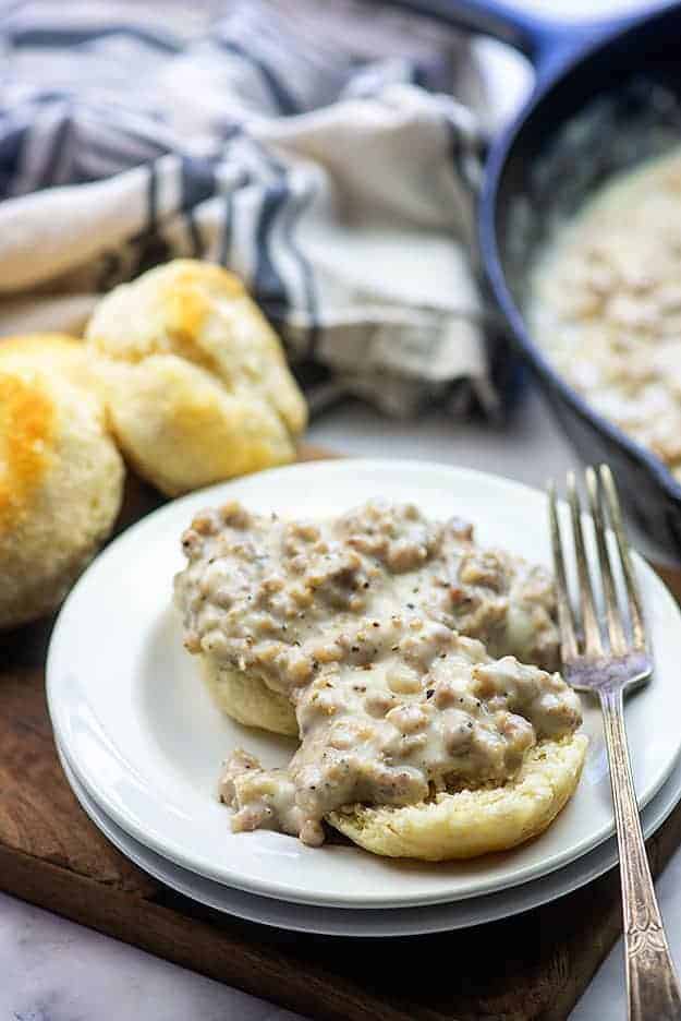 A single biscuit split in half topped with gravy in front of a cast-iron skillet.