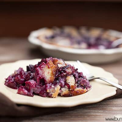 A serving of blackberry cobbler on a white plate in front of a cobbler pie.