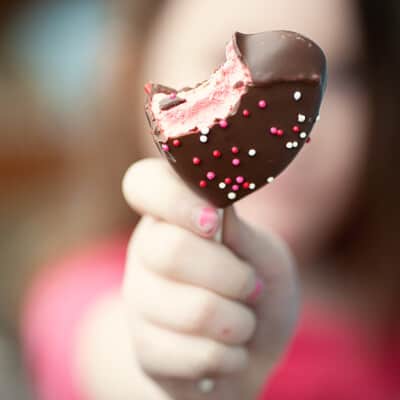 A girl holding up a chocolate heart shaped lollipop with a bite out of it.