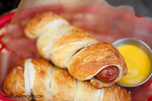 A closeup of a pretzel red hot dog over a cup of sauce 