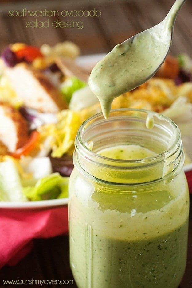 A close up of a spoon dripping salad dressing above a glass jar.
