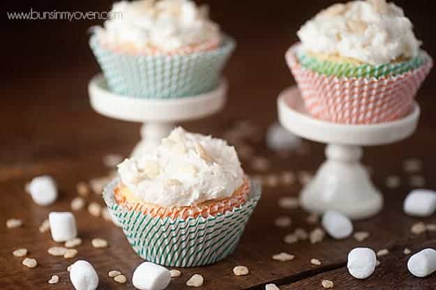 Three decorated cupcakes on a table surrounded by marshmallows and sprinkles.