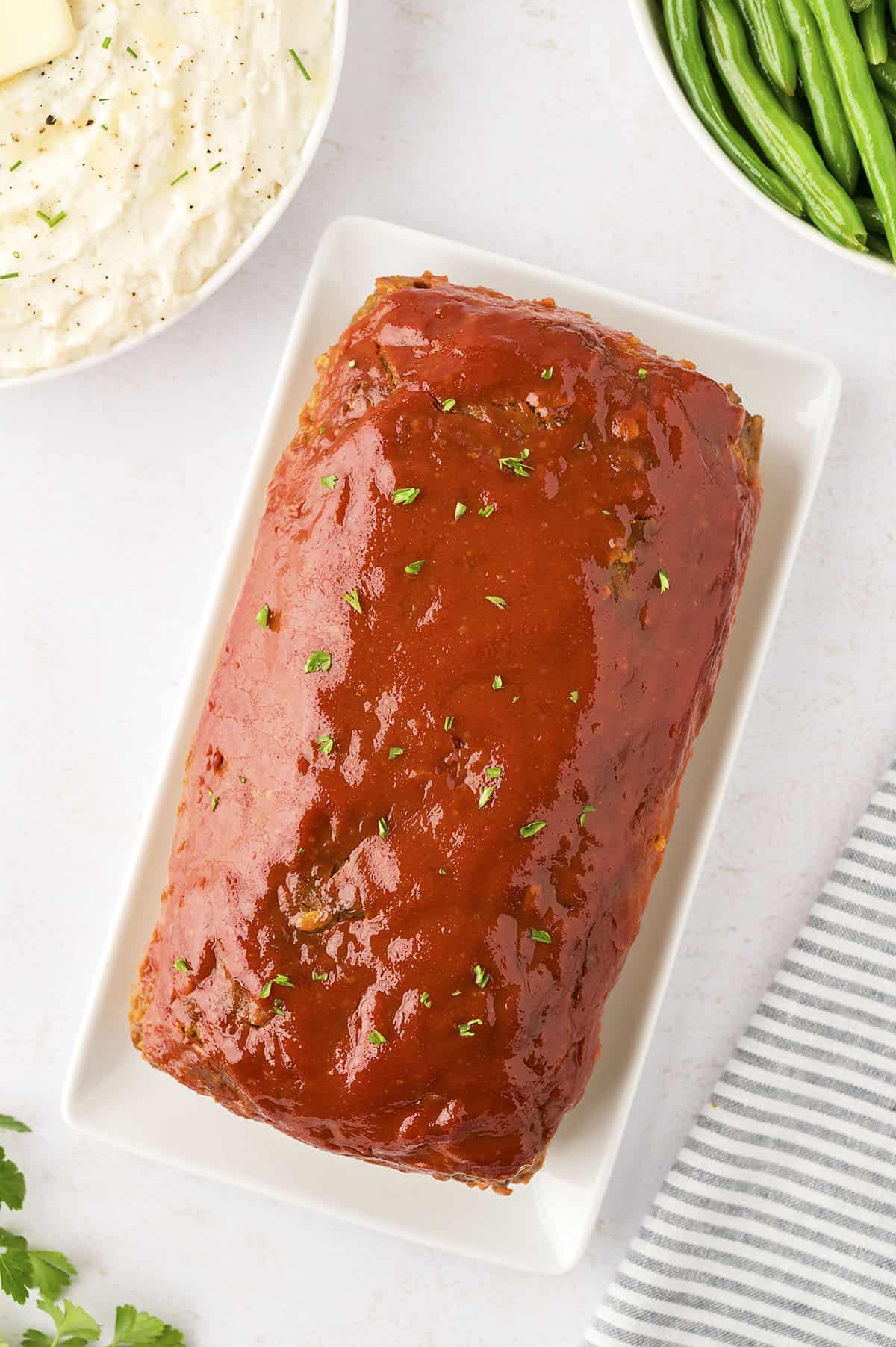 Saucy cheeseburger meatloaf on white platter next to mashed potatoes and green beans.