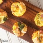 A narrow wooden cutting board with many ham strata muffins on it.