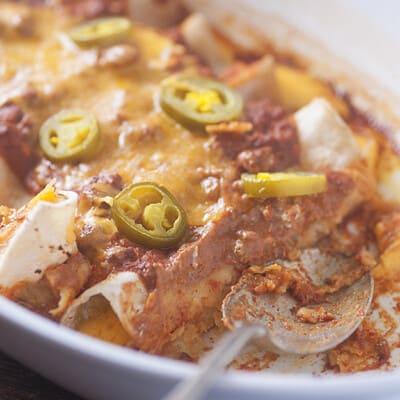 A spoon scooping out an enchilada from a white baking pan.