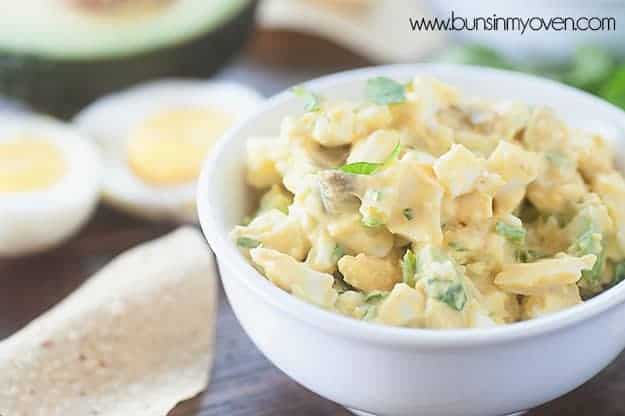 A close up of a white bowl of egg salad next to some chips on a table.