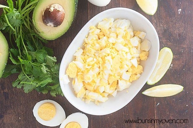 A bowl of egg salad on a table next to hard-boiled eggs and avocado.