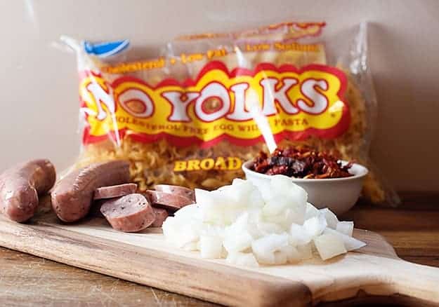 A bag of no yolks pasta behind onions and sausage on a wooden cutting board.