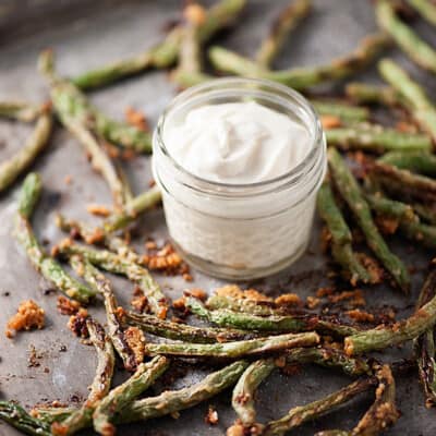 Parmesan coated green beans on a baking sheet with a jar of white dipping sauce.