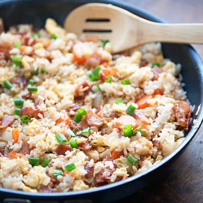 A close up of a skillet of bacon fried rice and a wooden spoon.
