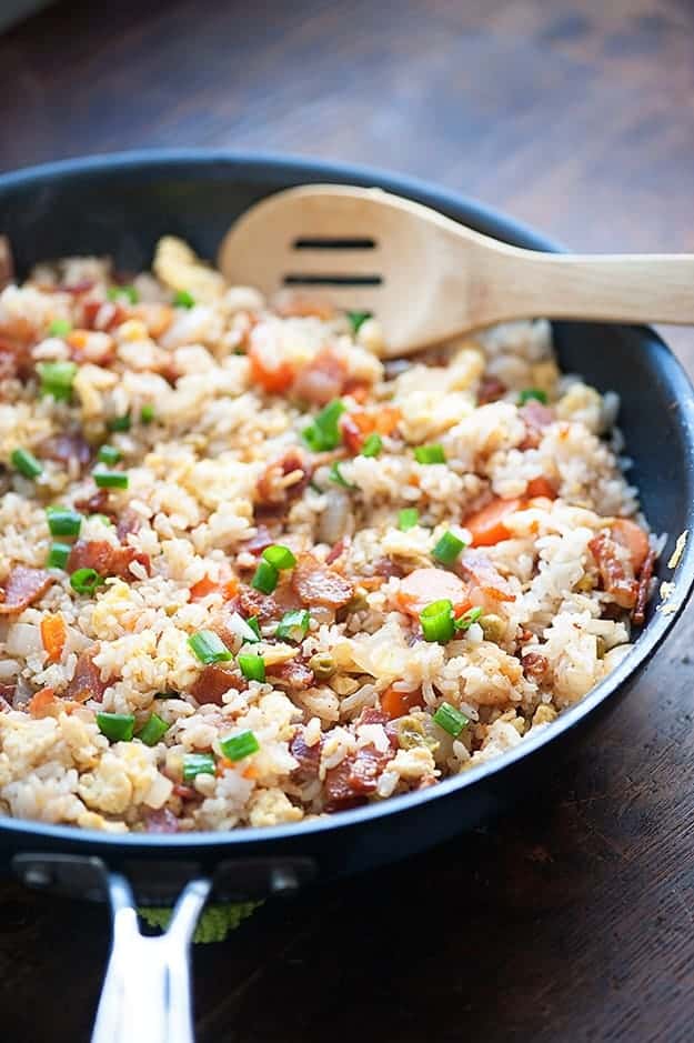 A close up of a skillet of bacon fried rice and a wooden spoon.