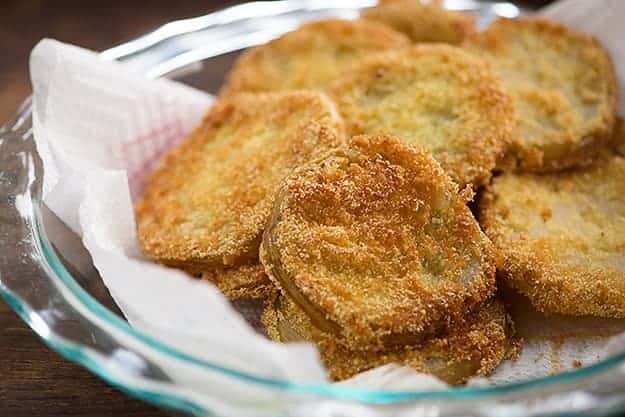 A clear glass pie plate lined with a paper towel with a pile of fried green tomatoes in it.