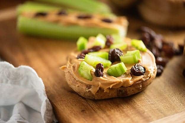 Celery, raisins, and peanut butter on top of mini bagels on a wooden cutting board.