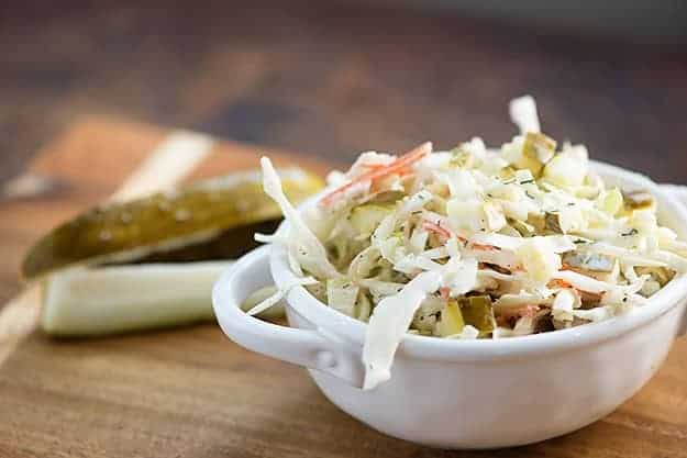 A bowl of coleslaw on a cutting board with a sliced pickle spear in the background.