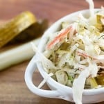 Close up of a bowl of cole slaw on a wooden cutting board in front of pickle spears.