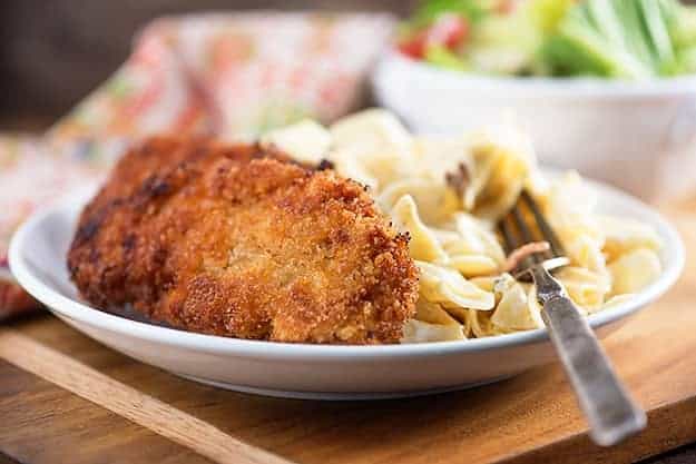 A close up of a breaded pork chop on a white plate atop a wooden cutting board.