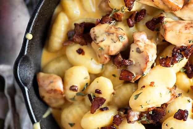 A close up of pork chunks and bacon in cheesy gnocchi in a cast-iron skillet.