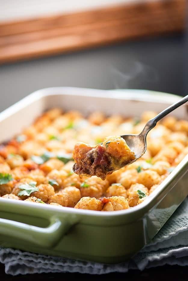 A spoon full of casserole in front of a baking pan full of tater tots.
