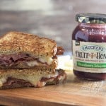 A close up of a sandwich sitting on top of a wooden table next to a jar of fruit spread.