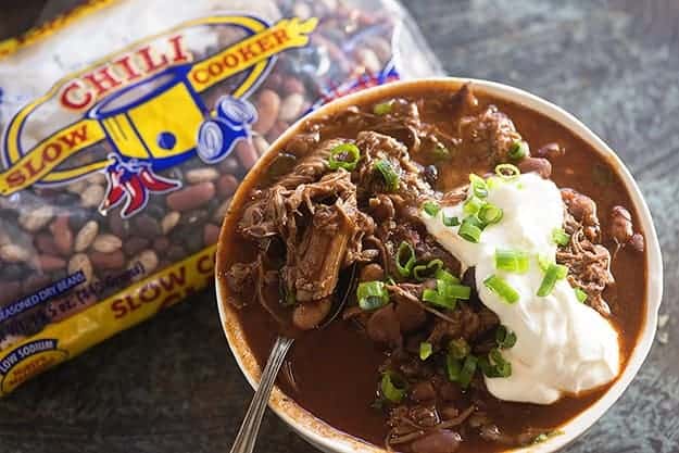 A bowl of chili topped with sour cream and chives in front of a bag of beans.