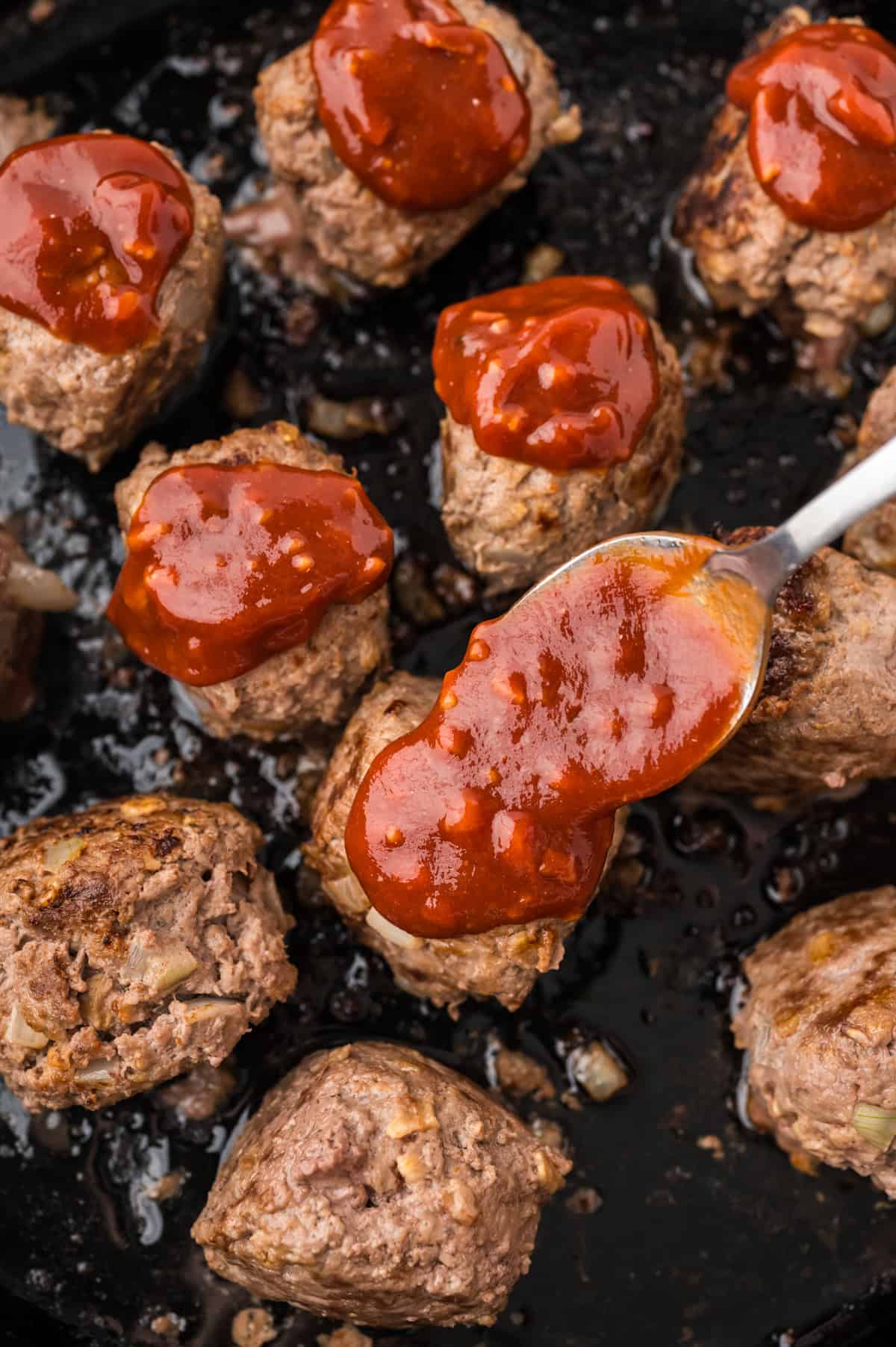 Spoon placing bbq sauce over meatballs in skillet.