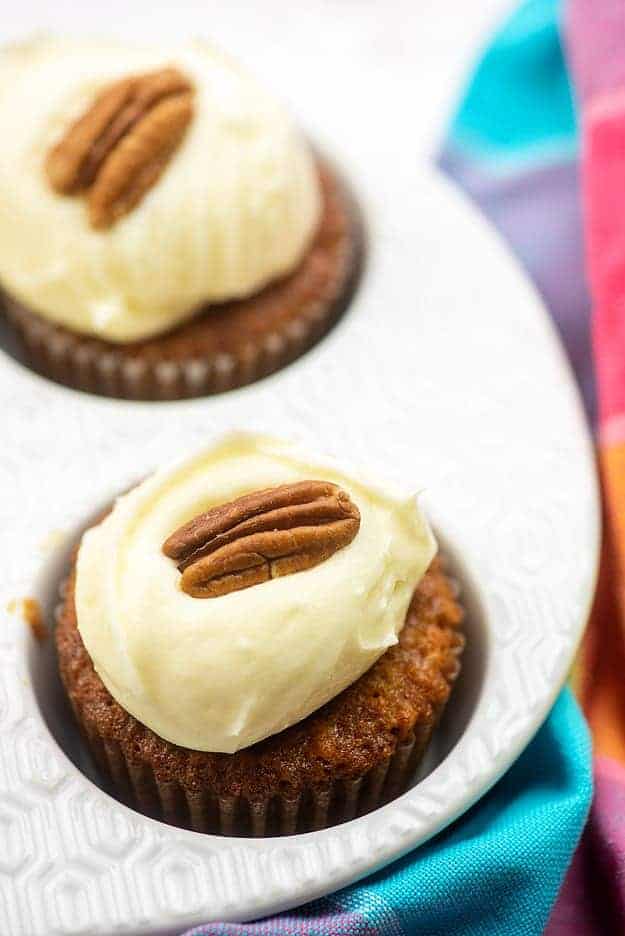 carrot cake cupcakes with cream cheese frosting and a pecan on top in white muffin tin