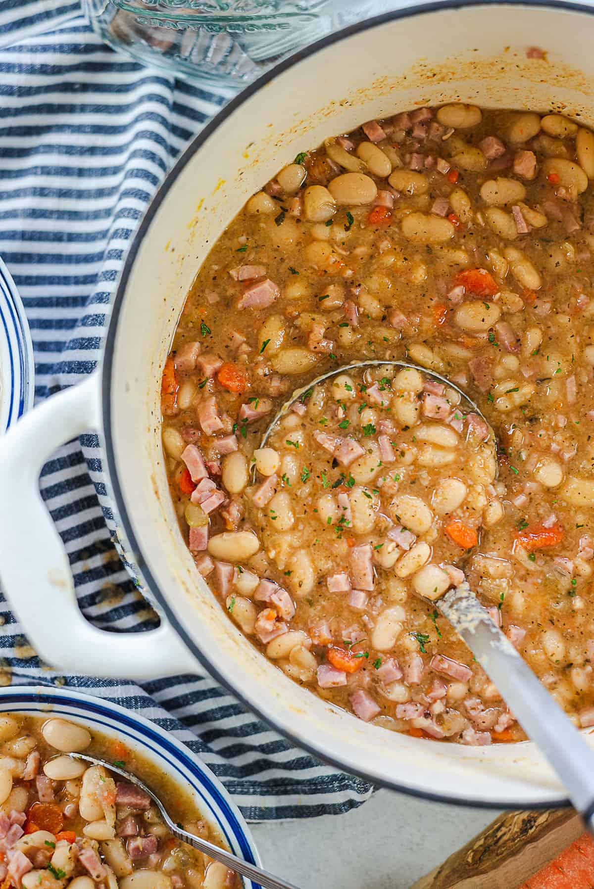 A ladle scooping white bean and ham soup from a large pot on a table next to bowls of soup.