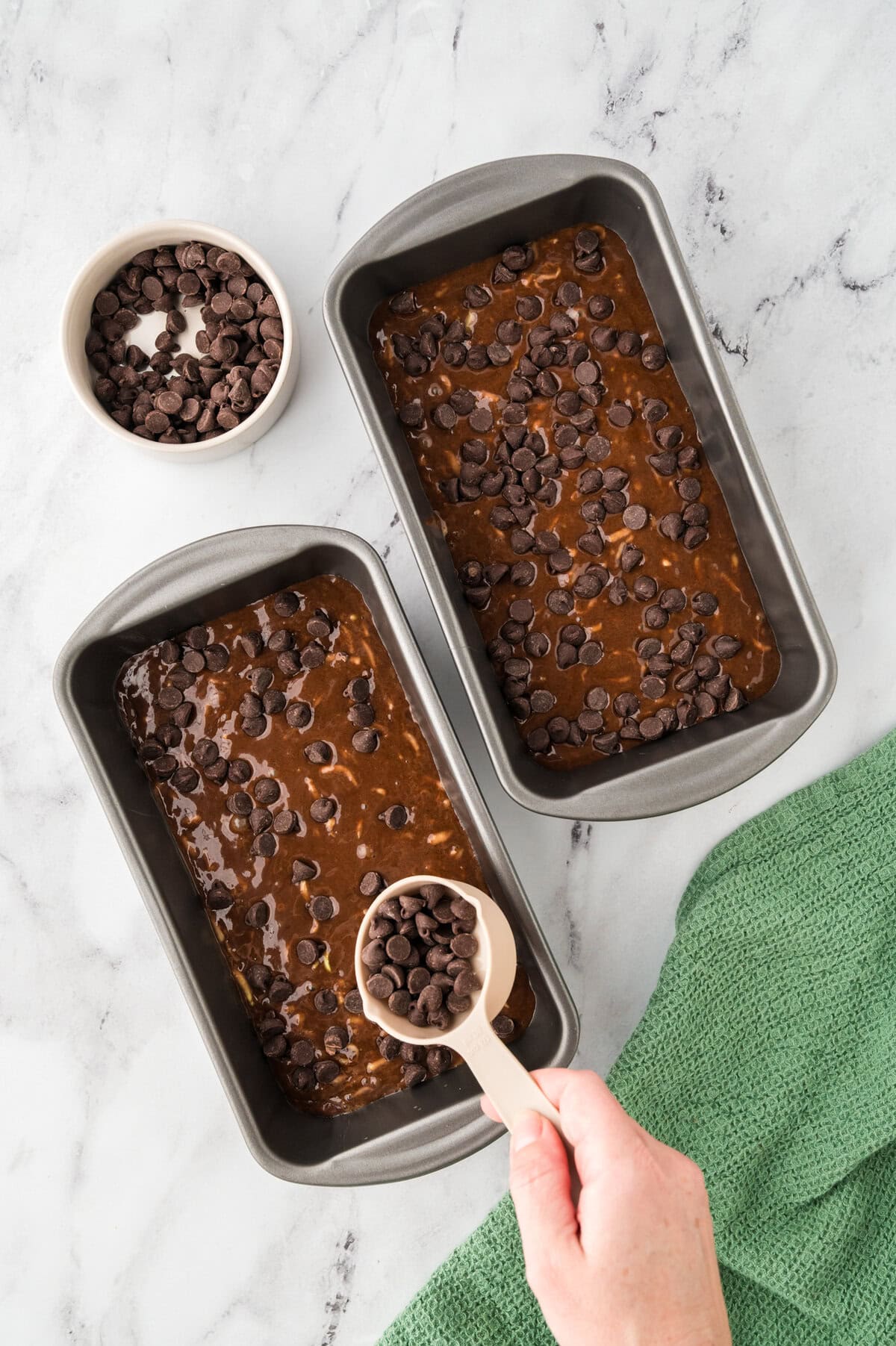 Chocolate chocolate chip zucchini bread batter in loaf pan being topped with chocolate chips.