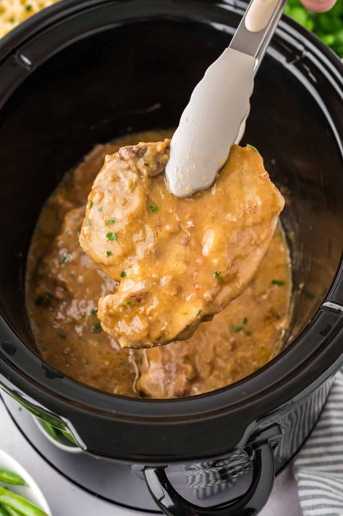 Smothered pork chops in crockpot on tongs.