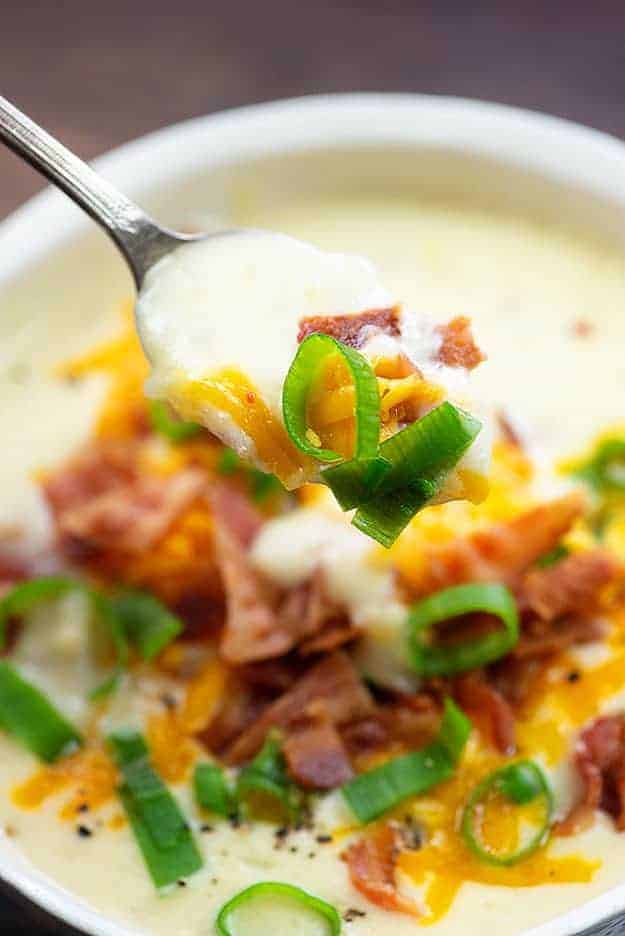 A close up of a spoon holding up potato soup over a bowl of soup.
