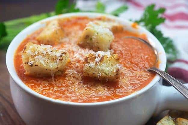 A white bowl of tomato soup with croutons on top 
