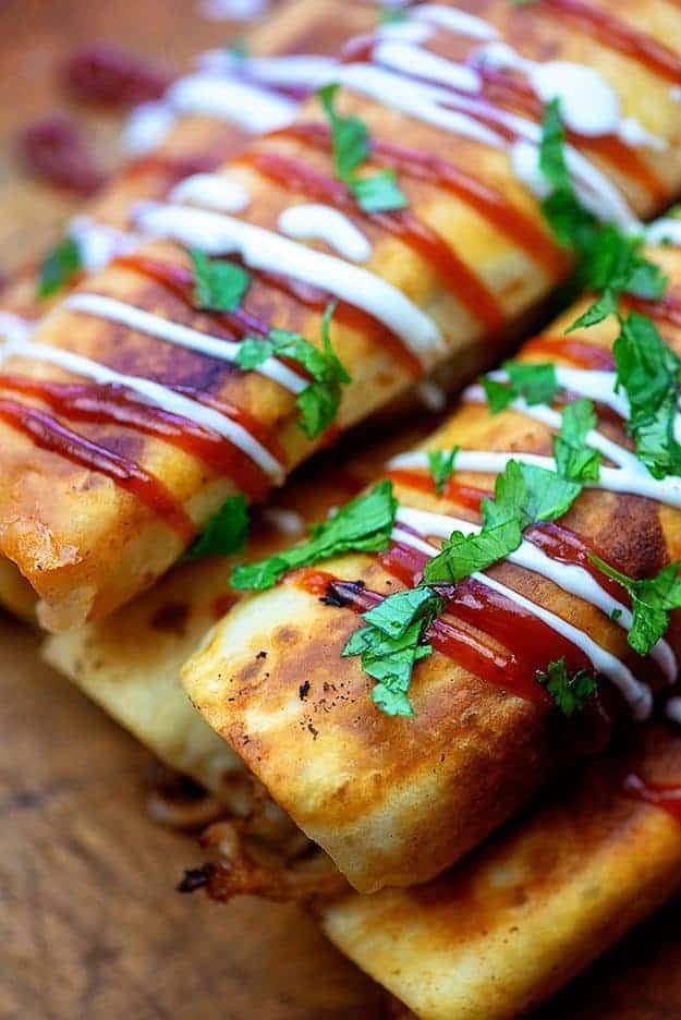 A close up of taquitos on a cutting board topped with drizzled ranch and bbq sauce.