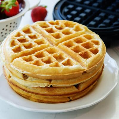 stack of belgian waffles on white plate next to waffle iron.