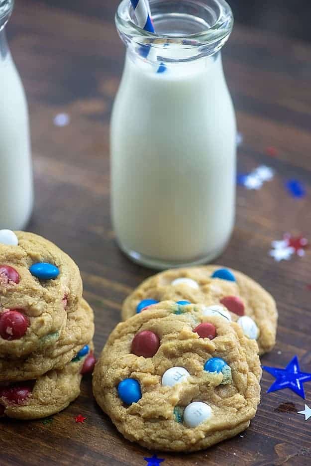 Several M&M cookies with a jar of milk on a table.