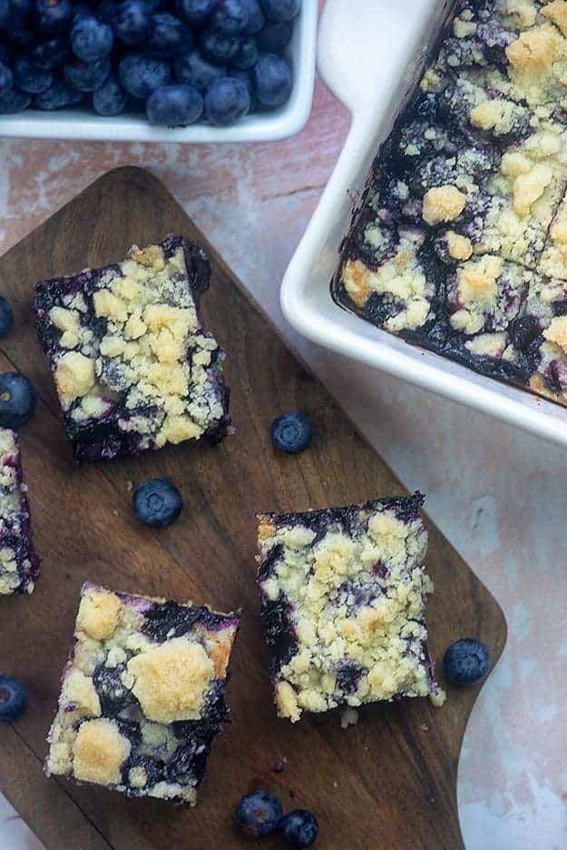 Individual blueberry bars on a cutting board in front of a baking pan with blueberry bars in it 