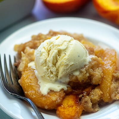 Peach cobbler topped with vanilla ice cream and a fork on a white plate