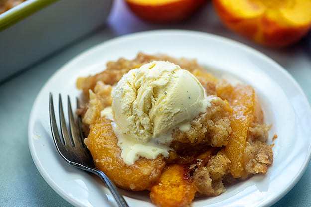 Peach cobbler topped with vanilla ice cream and a fork on a white plate