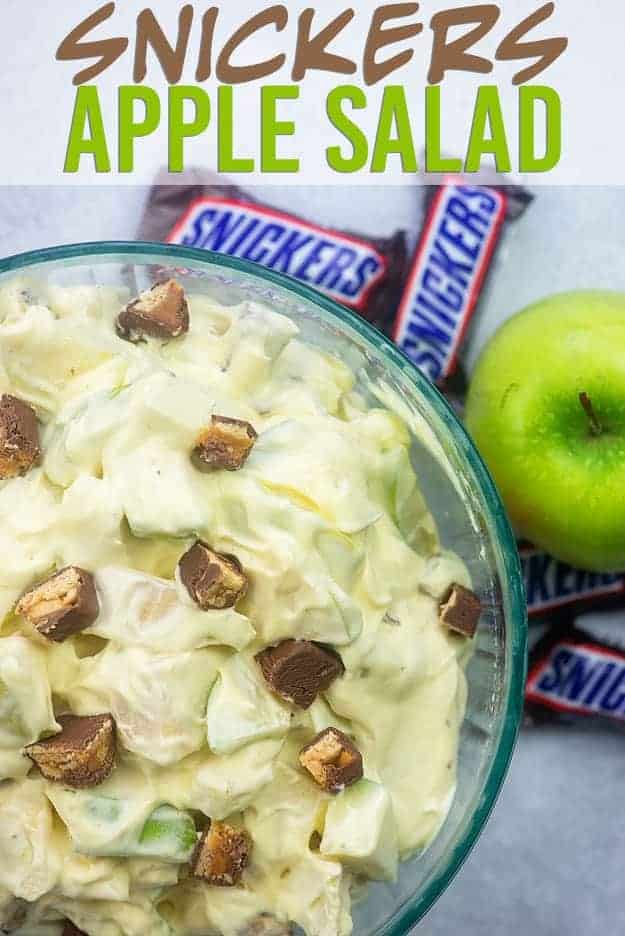 A clear glass bowl of snicker salad with whole apple and snickers in the background.