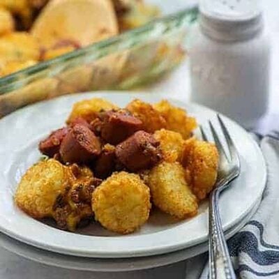 Round white plate with tater tots and pieces of hot dog on it.