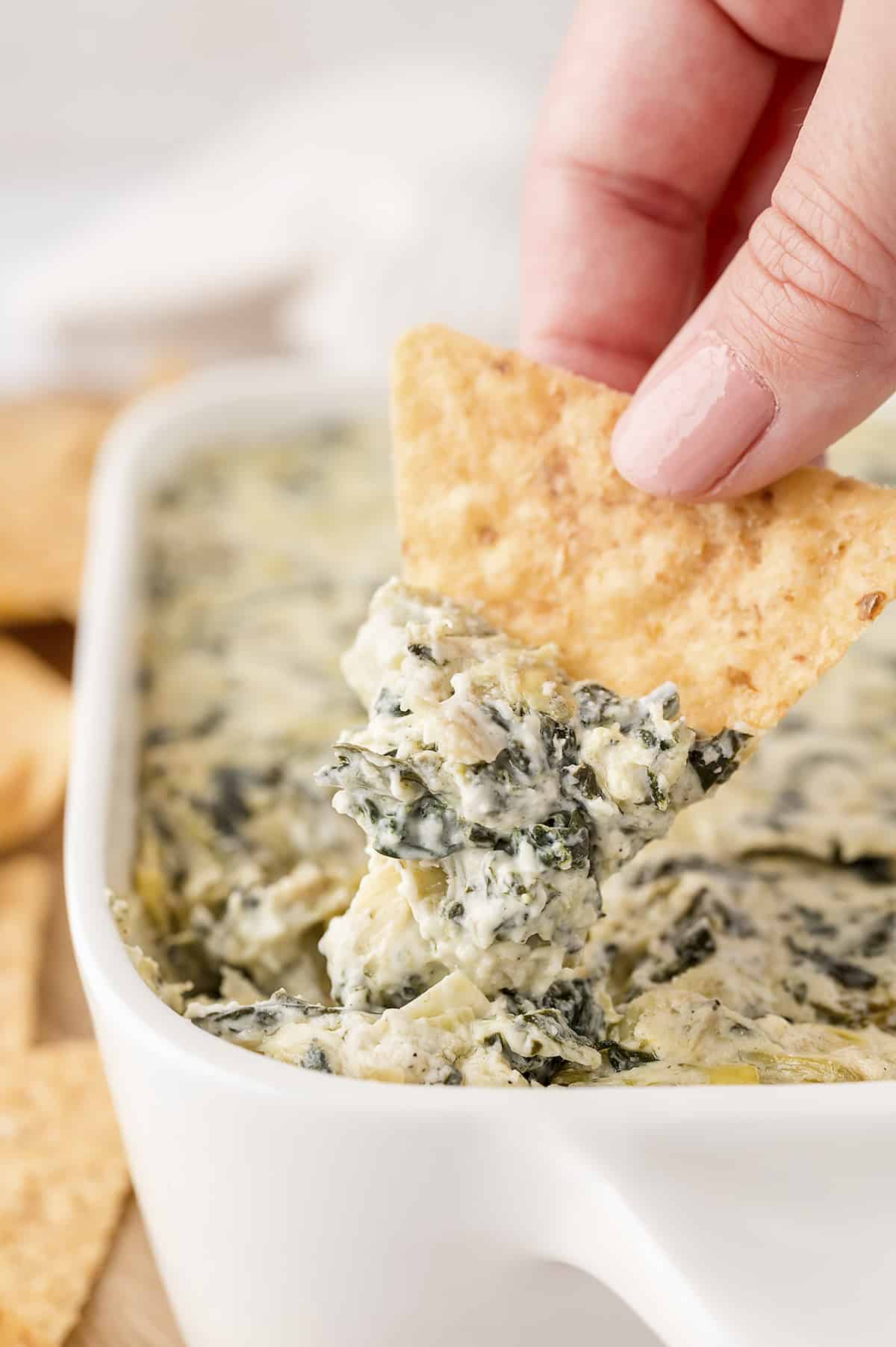 Hand dipping a tortilla chip into dish full of spinach artichoke dip.