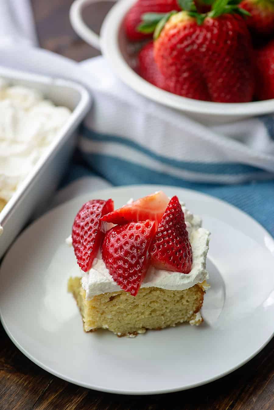 A slice of strawberry cream cake topped with whipped cream and fresh strawberries on a white plate.