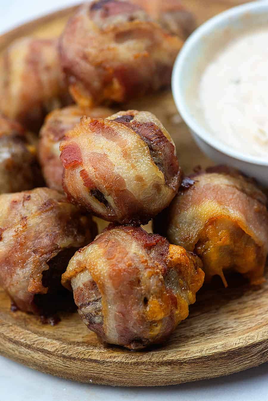 Bacon wrapped cheesburger bombs on a wooden tray next to a bowl of dipping sauce.