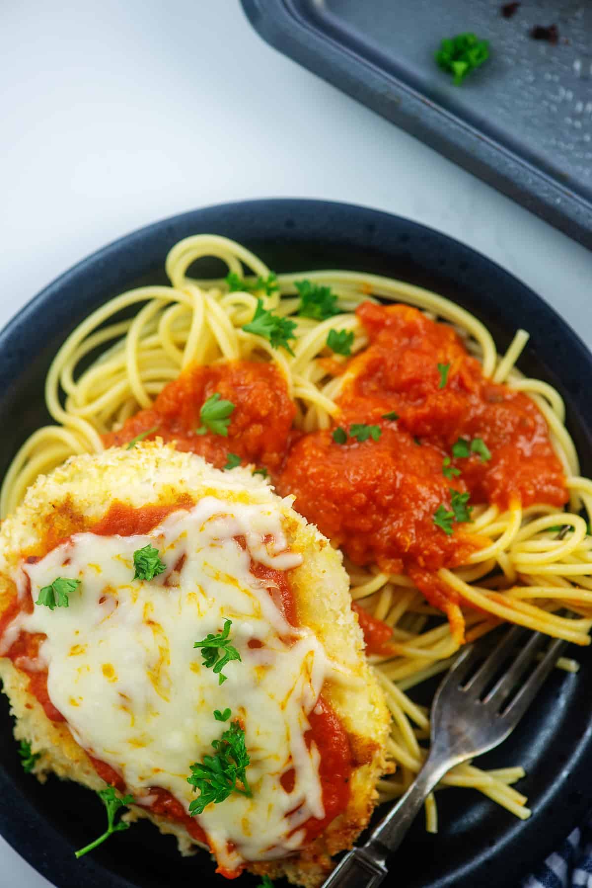 overhead view of baked chicken parmesan on black plate with a pile of spaghetti.