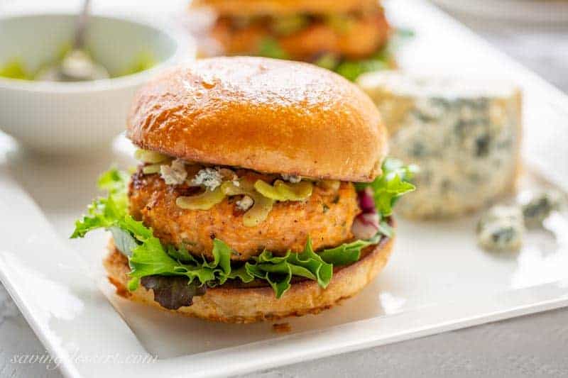 A close up view of a buffalo chicken burger with bun and condiments resting on a white plate.